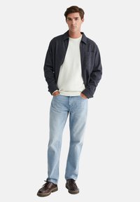 Donkergrijs colbert, licht getextureerde trui, lichtblauwe jeans, bruine schoenen. Model staat met handen in de zakken, neutrale achtergrond.