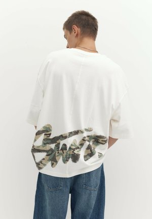 Jeune homme de dos portant un T-shirt blanc oversize avec un motif camouflage vert et marron et un jean bleu en denim.