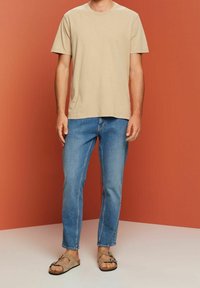T-shirt en coton beige, à manches courtes, associé à un jean en denim bleu clair et des sandales marron. Couleurs unies, coupe décontractée et détails minimalistes.
