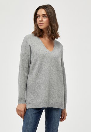 Pull en tricot gris avec un col en V, coupe décontractée et manches longues. Il a des poignets et un ourlet côtelés. Porté avec un jean en denim bleu.