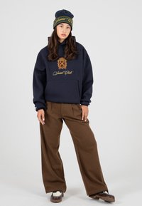 Granatowy hoodie z złotą grafiką i logo z przodu, w połączeniu z brązowymi spodniami o szerokich nogawkach i ciemną czapką z żółtym napisem.