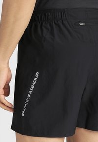 Henkilö, joka on pukeutunut mustiin Under Armour -shortseihin, joissa on joustava vyötärö ja vetoketjulla varustettu tasku takana oikealla puolella.