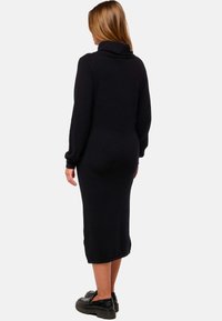 Robe en tricot noire avec un col drapé, des manches longues et une silhouette ajustée, se terminant au milieu du mollet. Portée avec des chaussures noires chunky.