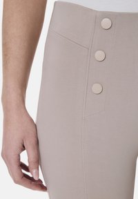 Beige hochtaillierte Hose mit glatter Textur, drei runden Knöpfen an der Seite und dezenten Nähten.