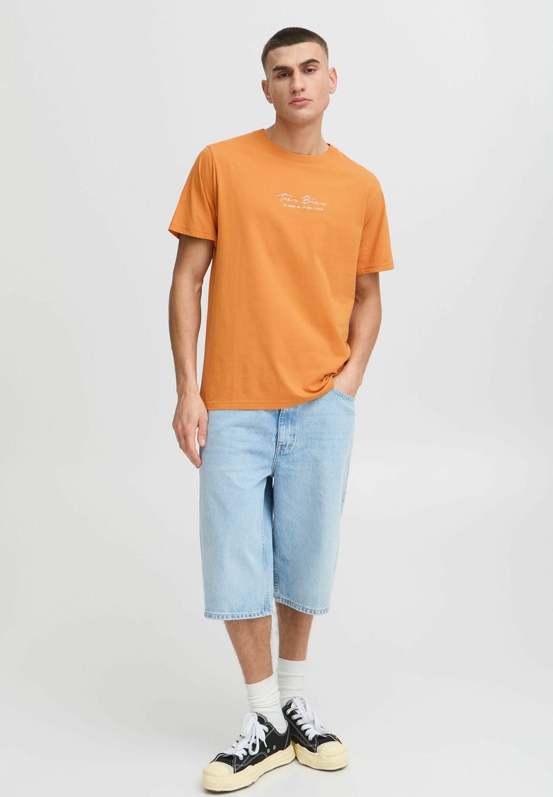 Orange bomulds t-shirt med korte ærmer og rund hals, parret med lyseblå denimshorts. Sorte sneakers med hvide snørebånd.
