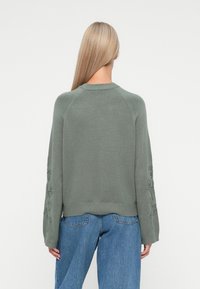 Pull vert en tricot avec des manches raglan, doté d'une broderie florale sur les bras, d'une finition côtelée et d'une coupe décontractée. Porté avec un jean bleu.