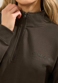 Felpa in pile con zip marrone, dotata di un colletto alto e logo in rilievo. Presenta un tessuto strutturato e una chiusura a zip a tre quarti sul collo.