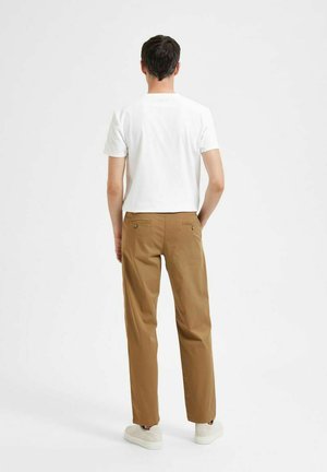Camisa blanca de manga corta combinada con pantalones rectos marrones con dos bolsillos traseros y zapatillas blancas, vista desde atrás.