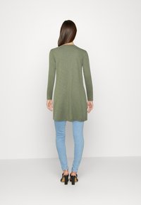 Túnica de manga larga verde oliva hecha de una tela suave, combinada con jeans ajustados azules claros y sandalias de tacón negras.