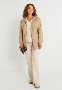 Femme souriante avec des cheveux bouclés portant une veste beige, un pull crème, un pantalon crème, des baskets beiges, tenant un sac à main en imprimé léopard.
