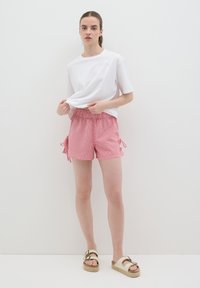 Pantalones cortos a cuadros rojos y blancos con lazos laterales, combinados con una camiseta blanca de corte holgado. El modelo está de pie sobre un fondo blanco con sandalias.