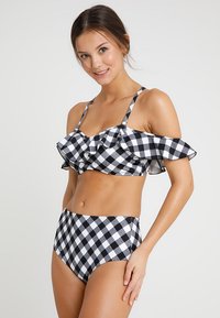 Biquíni em preto e branco com padrão xadrez, com parte de cima com babados e alças ajustáveis. Parte de baixo de cintura alta com o mesmo padrão xadrez.