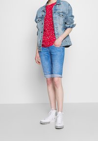 Chaqueta vaquera sobre una camiseta roja con estampado, combinada con pantalones cortos azul claro remangados y zapatillas altas blancas con detalles en negro.