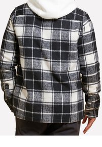 Chemise en flanelle à carreaux noir et blanc avec un tissu texturé ressemblant à de la laine, dotée d'une capuche blanche et de poignets boutonnés. Vue de dos montrant une coupe décontractée.