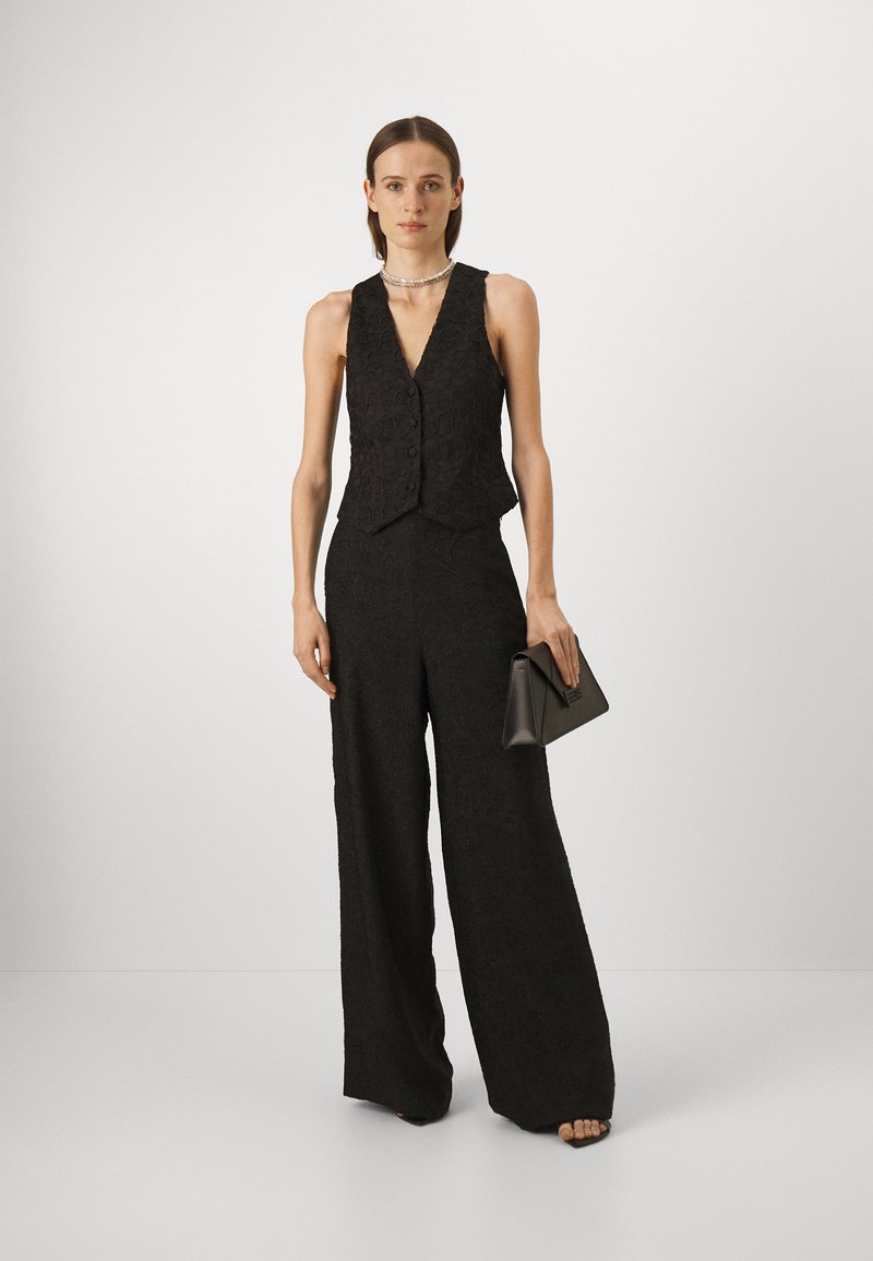 Schwarze, strukturierte Weste mit V-Ausschnitt und Knöpfen, kombiniert mit einer weiten Hose. Das Modell hält eine kleine schwarze Clutch. Neutrale Hintergrund.