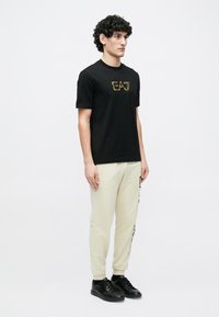 Görög hajú fiatal férfi áll jobbra nézve, fekete EA7 logós pólót, krémszínű oldalsó feliratos melegítőnadrágot és fekete sportcipőt visel.