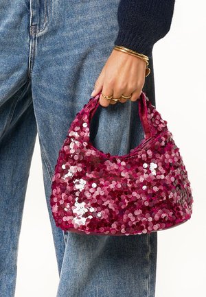 Sac à main rose pailleté et brillant, de forme arrondie, avec deux petites poignées, tenues dans une main sur un tissu en denim.