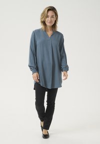 Bluza s dolgimi rokavi v teal barvi z V-izrezom in diamantsko teksturo, v kombinaciji s ozkimi črnimi hlačami in črnimi balerinkami. Preprost, sproščen dizajn.