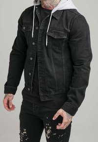 Svart denimjacka med grå huva, med knäppning framtill, två bröstfickor och en slim fit-design. Bärs över en svart T-shirt och slitna jeans.