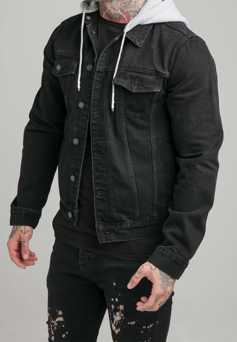 Svart denimjacka med grå huva, med knäppning framtill, två bröstfickor och en slim fit-design. Bärs över en svart T-shirt och slitna jeans.