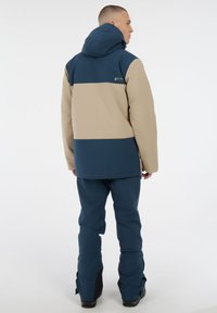 Blaue und beige Jacke mit Kapuze, die ein Farbblockdesign aufweist. Kombiniert mit passenden blauen Hosen und schwarzen Akzenten am unteren Saum.