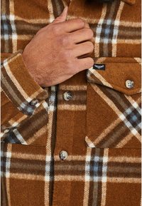 Braunes Fleecehemd mit kariertem Muster, das weiße, hellblaue und braune Akzente aufweist, mit Knopfverschlüssen und Brusttaschen.