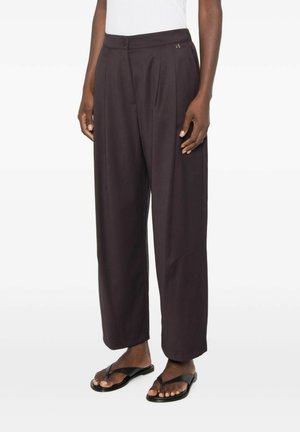 Persona che indossa pantaloni larghi marrone scuro con vita abbottonata e sandali neri aperti, in piedi davanti a uno sfondo semplice.