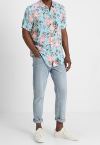 Chemise à manches courtes, de couleur bleu clair, ornée de motifs floraux roses et verts, associée à un jean en denim clair et à des baskets blanches.