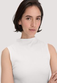Witte mouwloze top met een hoge halslijn, voorzien van een geribbelde textuur en gladde stof. Eenvoudig ontwerp, aansluitende snit en minimale details.