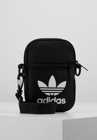 Czarna torba crossbody Adidas z białym logo trefoil, frontową kieszenią na zamek i regulowanym paskiem, na białej powierzchni i jasnoszarym tle.