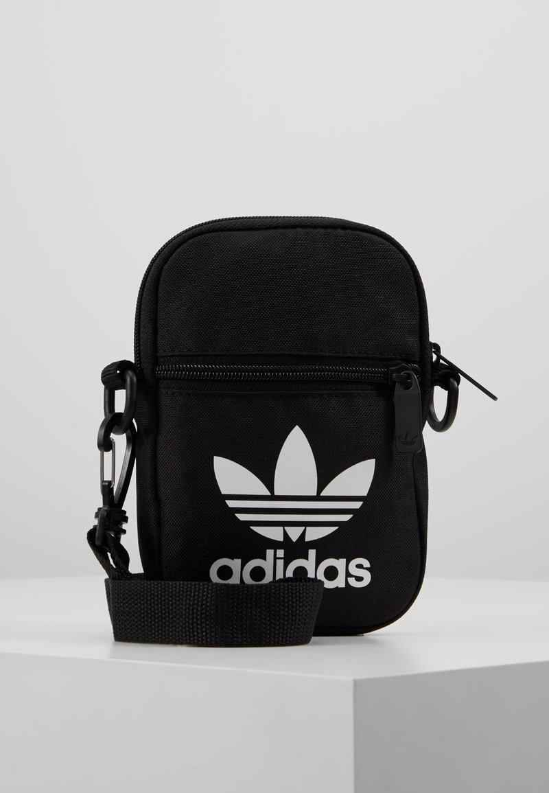 Czarna torba crossbody Adidas z białym logo trefoil, frontową kieszenią na zamek i regulowanym paskiem, na białej powierzchni i jasnoszarym tle.