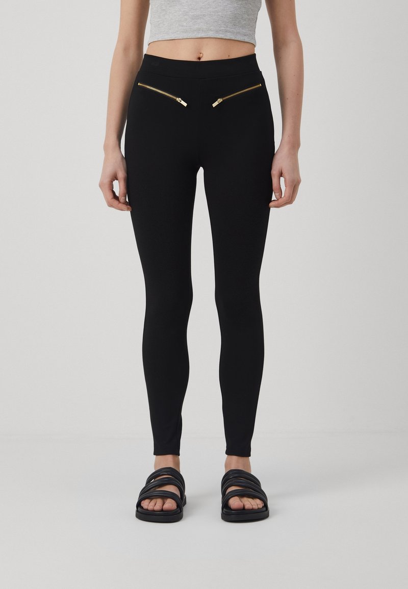 Leggings noirs au toucher lisse, arborant deux poches zippées dorées à l'avant et une taille haute. Associés à des sandales noires.