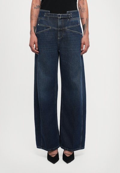 Jean en denim bleu à jambes larges et taille haute, avec passants de ceinture, deux poches avant et des détails de couture contrastés.