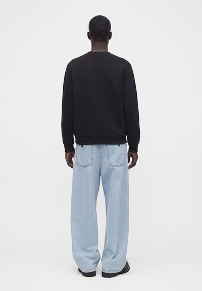 Homme de dos portant une chemise noire à manches longues, un jean large bleu clair et des chaussures noires, debout devant un fond clair uni.