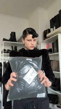 Black pillow packaged in a clear plastic bag, with a white label attached. Background features black footwear and accessories on shelves.