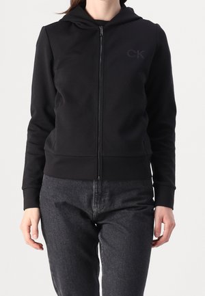 Kobieta w czarnej bluzie z kapturem zapinanej na zamek błyskawiczny z logo "CK" na piersi i ciemnoszarych dżinsach, stojąca na jednolitym jasnym tle.