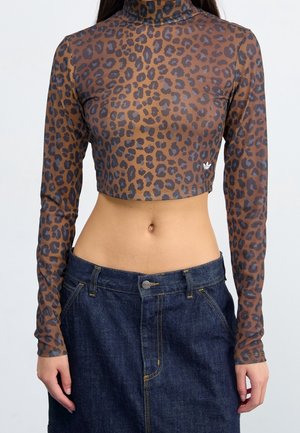 Frau trägt ein braunes Crop-Top mit Leopardenmuster und langen Ärmeln sowie einen dunkelblauen, hoch taillierten Jeansrock, steht vor einem einfarbigen Hintergrund.