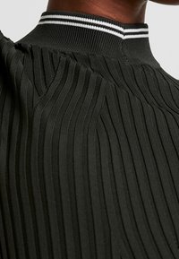 Zbliżenie czarnego swetra w serek z prążkowanej tkaniny, z białymi podwójnymi paskami na kołnierzu, noszonego przez osobę.