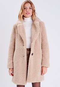 Cappotto sherpa beige con una texture soffice, ampio risvolto, chiusura con un singolo bottone e maniche lunghe, indossato sopra un maglione bianco di maglia e una gonna marrone.