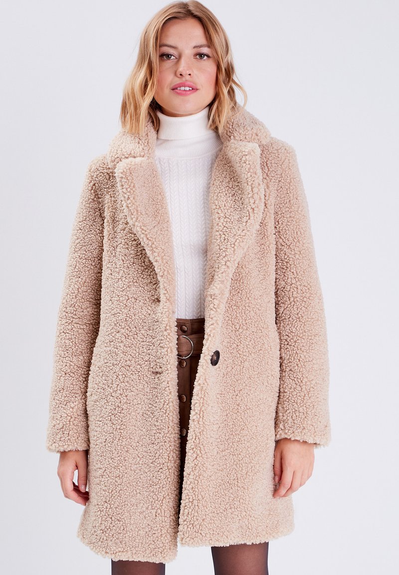Cappotto sherpa beige con una texture soffice, ampio risvolto, chiusura con un singolo bottone e maniche lunghe, indossato sopra un maglione bianco di maglia e una gonna marrone.