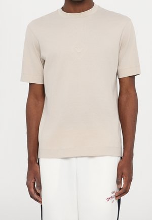 Béžové tričko s krátkým rukávem vyrobené z měkké tkaniny, se středovým malým logem ve tvaru diamantu. Kulatý výstřih a uvolněný střih.