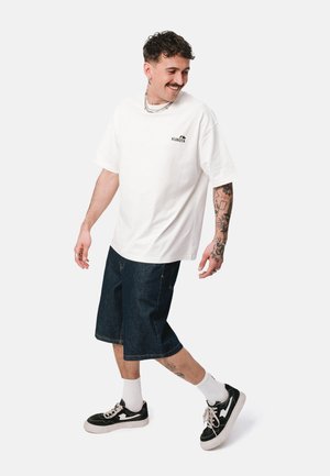 Mężczyzna z tatuażami, ubrany w białą koszulkę Kubota, dżinsowe szorty, białe skarpetki i czarne trampki, stoi i uśmiecha się patrząc w dół.