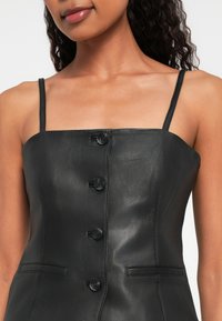 Naine kannab musta nahast ilma varrukateta pintsakut, millel on õhukesed rihmad ja neli nuppu ees, avades õlad ja ülemise rinna.