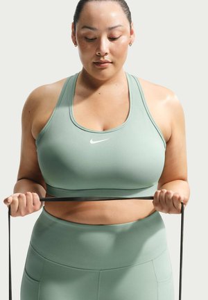 Kobieta w jasnozielonym sportowym biustonoszu i legginsach trzymająca taśmę oporową, patrząca w dół, skoncentrowana, na jednolitym tle.