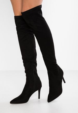 Bottes montantes en daim noir avec bouts pointus, fermetures éclair latérales et talons hauts fins, portées sur des jambes croisées sur un fond blanc.