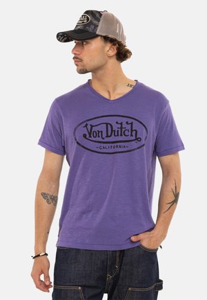 Homme portant un t-shirt violet Von Dutch California, une casquette trucker noire et beige, un jean foncé, montrant des tatouages sur les deux bras, regardant sur le côté.