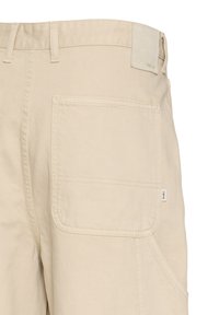 Pantalon beige avec grande poche carrée à l'arrière, passants de ceinture, et petite étiquette de marque sur le bord de la poche.