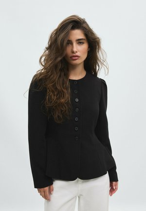 Femme aux longs cheveux bruns ondulés portant une veste noire cintrée boutonnée et un pantalon blanc sur un fond clair uni.