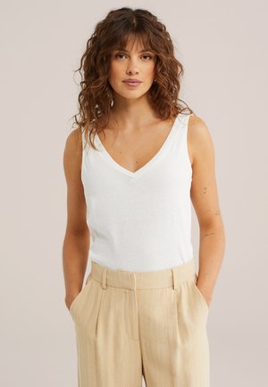 Femeie cu păr creț brunet, purtând un top alb fără mâneci cu decolteu în V și pantaloni bej cu dungi fine, cu mâinile în buzunare, fundal neutru.