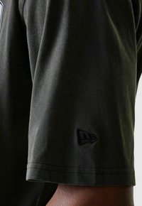 Gros plan sur un t-shirt noir à manches courtes porté sur un bras, montrant un logo brodé subtil près de l'ourlet de la manche.